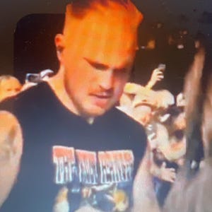 May include: A person wearing a black sleeveless shirt with the text "THE FINAL FESTIVAL" in white and red. The person has short, light-colored hair and is in a crowd of people.