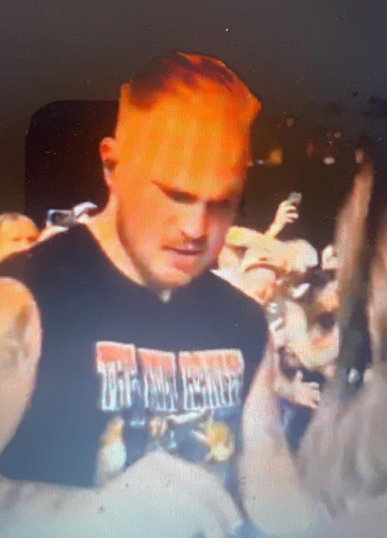 May include: A person wearing a black sleeveless shirt with the text "THE FINAL FESTIVAL" in white and red. The person has short, light-colored hair and is in a crowd of people.
