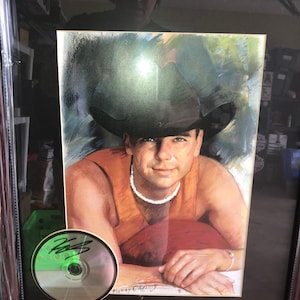 May include: Framed portrait of a man in a cowboy hat and orange tank top, with a CD featuring an autograph. The portrait is in a dark brown frame, and the CD is signed by Kenny Chesney.