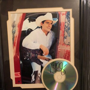 May include: Framed photograph of a man in a white cowboy hat and shirt, holding a guitar. A CD with an autograph is in front of the photo. The frame is black with a white mat.