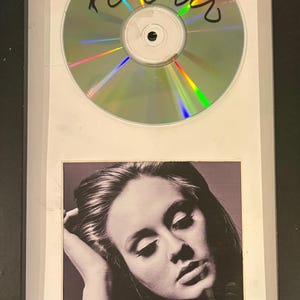Puede incluir: Un marco que exhibe un disco compacto firmado con una autógrafo en tinta negra sobre la superficie reflectante, colocado sobre la portada del álbum Adele 21, que muestra un retrato en blanco y negro de la artista con el texto ADELE 21.