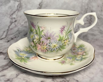 May include: A white teacup and saucer with a floral pattern. The cup has a gold rim and the saucer has a gold trim. The flowers are pink, purple, and blue.