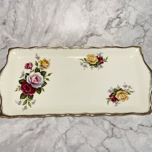 Può includere: Un vassoio rettangolare bianco per servire con un bordo dorato e disegni floreali. Il vassoio presenta tre gruppi di rose in tonalità di rosa, rosso e giallo con foglie verdi e dettagli blu.