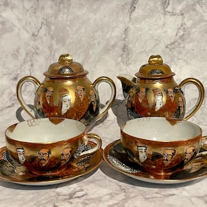 Op de afbeelding: Een vintage Japanse theeset met gouden accenten en ingewikkelde ontwerpen. De set bevat een theepot, een suikerpot en twee theekopjes met schoteltjes. De theekopjes en schoteltjes hebben een zwart en gouden ontwerp met een herhalend patroon van figuren.
