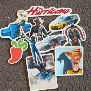 May include: A collection of die-cut stickers featuring race cars, drivers, and the text "The Hurricane." The stickers showcase various racing scenes, including cars in motion and drivers celebrating victories. The stickers are on a textured surface.