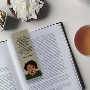 May include: A close-up of an open book with a bookmark featuring a woman's face and the text "We will all, at some point, encounter hurdles and barriers to gaining access to entry, but we can help moving up and overcoming those hurdles, but on the other side of the captivity is our own story and our own story."  The bookmark is on a page with text about places and adventure.