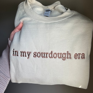 May include: A beige sweatshirt with the text "in my sourdough era" embroidered in brown on the front.