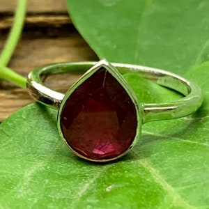 May include: A silver ring featuring a teardrop-shaped ruby gemstone. The ring is resting on a green leaf.