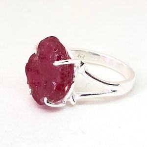 Op de afbeelding: Een zilveren ring met een grote, ruwe, rode edelsteen. De ring heeft een eenvoudig, open bandontwerp.