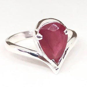 May include: A silver ring featuring a pear-shaped, deep red gemstone. The gemstone is set in a bezel setting, and the band has a simple, elegant design. The ring is displayed against a white background, highlighting its color and shape.