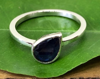May include: A silver ring with a teardrop-shaped, dark blue gemstone. The ring is set on a thin band and rests on a green leaf. The gemstone has a faceted surface, catching the light. The ring is displayed on a wooden surface.