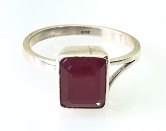 Könnte beinhalten: Ein silberner Ring mit einem rechteckigen, tiefroten Edelstein. Das Ringband ist dünn und schlicht, mit einem geteilten Design in der Nähe des Edelsteins. Der Edelstein ist in einer quadratischen Lünette gefasst. Der Ring befindet sich auf weißem Hintergrund.