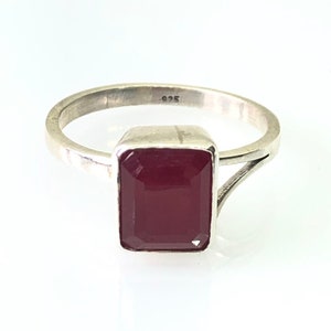 May include: A silver ring with a rectangular, deep red gemstone. The ring band is thin and simple, with a split design near the gemstone. The gemstone is set in a square bezel. The ring is on a white background.