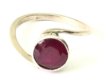 Puede incluir: Un anillo de plata con una piedra preciosa de rubí rojo. El anillo tiene un diseño simple y moderno con una banda curva.