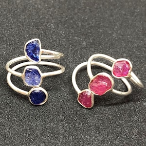 Peut inclure: Deux bagues en argent avec trois pierres précieuses chacune. La bague à gauche a trois saphirs bleus, et la bague à droite a trois rubis rouges.