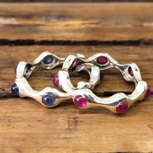 May include: Two silver rings with alternating blue and pink gemstones. The rings are interlocked and have a wavy design.