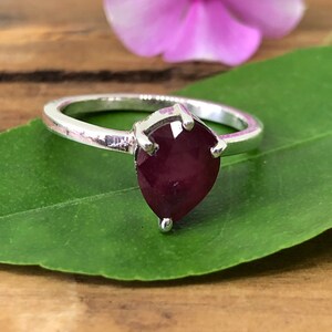 Peut inclure: Une bague en argent avec une pierre précieuse en rubis de forme poire. La bague est posée sur une feuille verte.