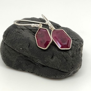 May include: A pair of silver earrings with a geometric design. Each earring features a deep red gemstone set in a silver frame. The earrings are displayed on a dark gray, textured surface against a white background.
