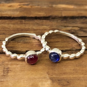 Könnte beinhalten: Zwei silberne Ringe mit perlenbesetzten Kanten, jeder mit einem runden Edelstein besetzt. Ein Ring hat einen roten Edelstein, der andere einen blauen Edelstein.