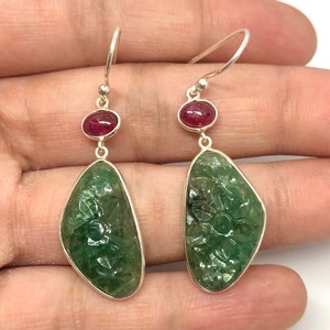 Może przedstawiać: Para kolczyków wiszących z zielonymi, rzeźbionymi kamieniami szlachetnymi i czerwonymi akcentami z kamieni szlachetnych.