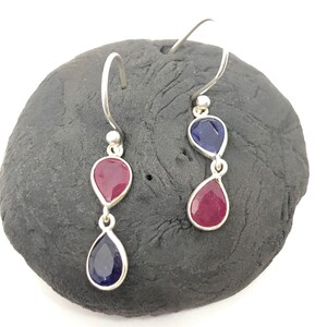 May include: A pair of silver drop earrings featuring a teardrop design. Each earring has two gemstones: a deep red and a dark blue, set in a silver frame. The earrings are displayed on a dark gray stone.