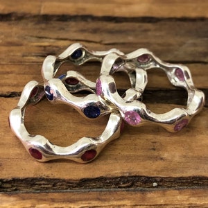 Könnte beinhalten: Drei silberne Ringe mit wellenförmigem Design und bunten Edelsteinen. Die Ringe sind übereinander gestapelt und haben rosa, blaue und rote Edelsteine.