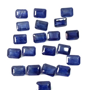 May include: A collection of 17 blue sapphire gemstones, cut in a rectangular emerald shape. The gemstones are arranged in a scattered pattern on a white background.