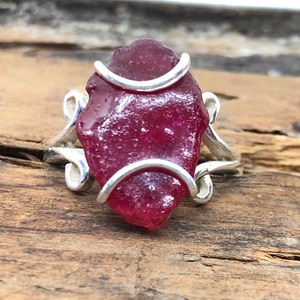 May include: A silver ring featuring a large, deep red, irregularly shaped piece of sea glass set in the centre. The ring is crafted with a delicate, twisted silver band.