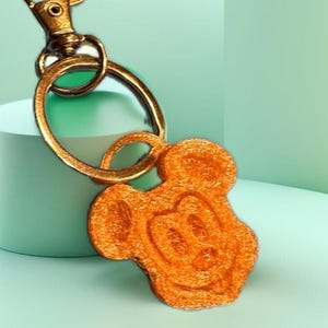 May include: Orange Mickey Mouse keychain with a gold-colored clasp and ring. The Mickey Mouse charm has a textured surface and is attached to a gold-colored metal clasp. The background is a light green color.