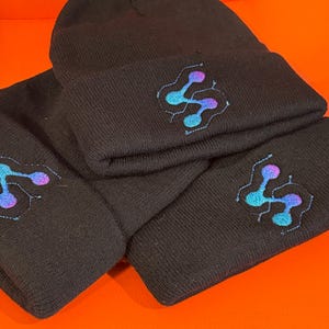 May include: Black knit beanie hats with a folded brim. Each hat features an embroidered design in shades of blue, purple, and teal. The hats are displayed on an orange surface.