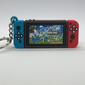 May include: A brown and black Nintendo Switch keychain with a blue and red controller on each side. The screen shows a video game with the text "SUPER MARIO BROS. U".