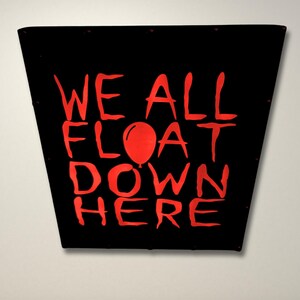 May include: Black and red sign with the text "WE ALL FLOAT DOWN HERE" in red lettering. A red balloon is in the middle of the text.