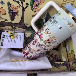 K&ouml;nnte beinhalten: Ein Stanley Quencher H2.0 Becher mit Winnie Puuh-Design, hellblauem Deckel und Griff. Der Becher befindet sich in einer dekorativen Box mit Winnie Puuh-Illustrationen, einer kleinen Bienenfigur und einem wei&szlig;en Kordelzugbeutel.