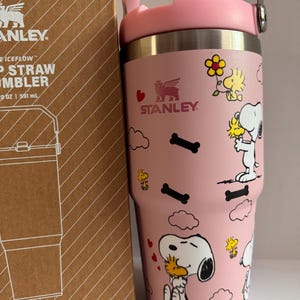 May include: A pink Stanley Iceflow straw tumbler with a pink handle and lid. The tumbler features illustrations of Snoopy and Woodstock, along with bone and cloud designs. The tumbler is next to a brown cardboard box with the Stanley logo and product information.