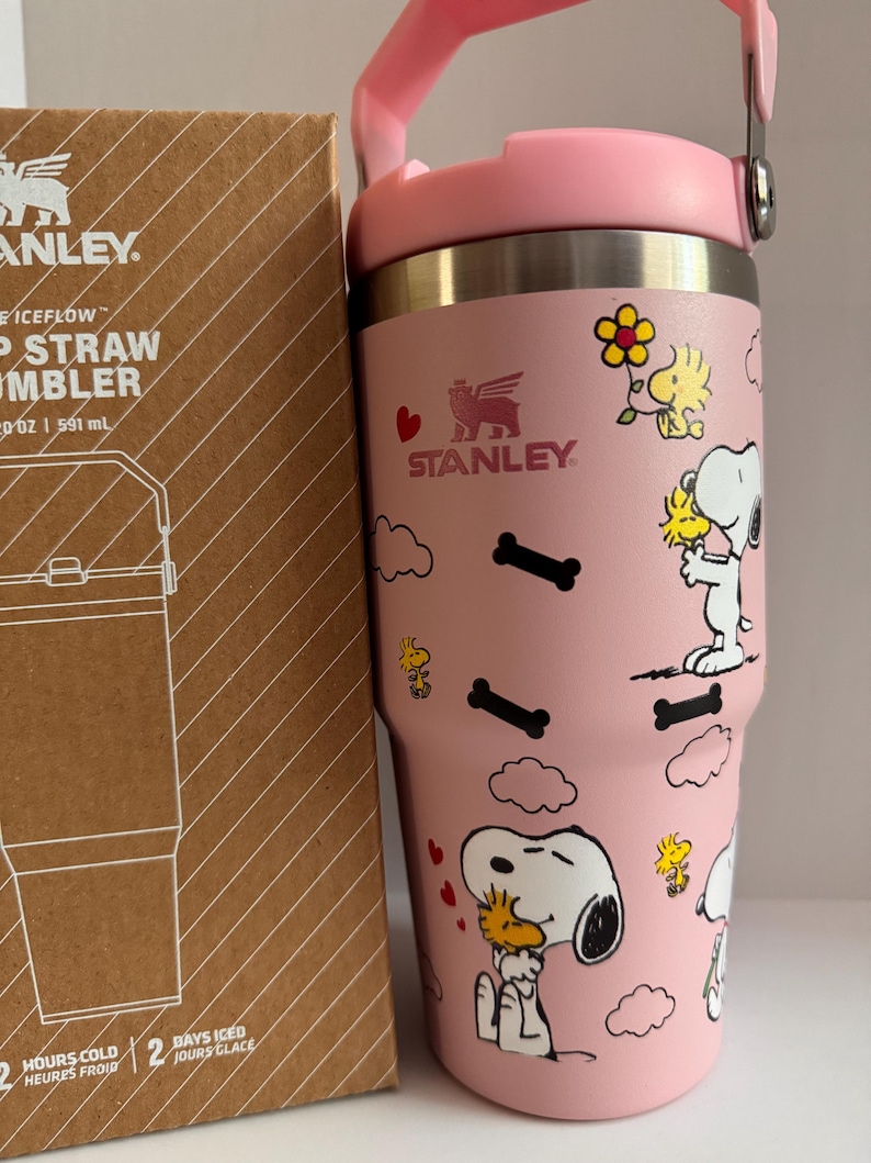 May include: A pink Stanley Iceflow straw tumbler with a pink handle and lid. The tumbler features illustrations of Snoopy and Woodstock, along with bone and cloud designs. The tumbler is next to a brown cardboard box with the Stanley logo and product information.