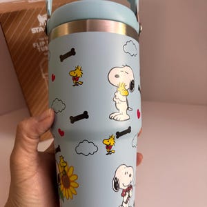 May include: A light blue tumbler with a handle, featuring Snoopy and Woodstock cartoon characters, bones, clouds, and hearts. The tumbler has a silver rim and a light blue handle. The tumbler is cylindrical and appears to be made of stainless steel.
