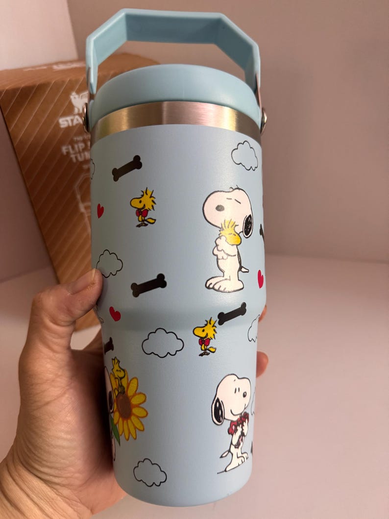 May include: A light blue tumbler with a handle, featuring Snoopy and Woodstock cartoon characters, bones, clouds, and hearts. The tumbler has a silver rim and a light blue handle. The tumbler is cylindrical and appears to be made of stainless steel.