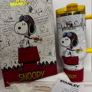 May include: A white Stanley tumbler featuring a Snoopy graphic in aviator gear, a yellow lid, and a yellow handle. The tumbler is displayed next to its matching box, which shows a comic strip pattern, and a white care and use instruction manual.