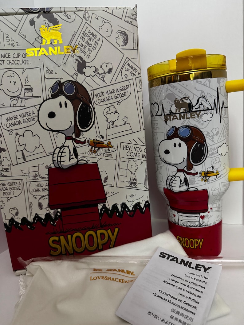 May include: A white Stanley tumbler featuring a Snoopy graphic in aviator gear, a yellow lid, and a yellow handle. The tumbler is displayed next to its matching box, which shows a comic strip pattern, and a white care and use instruction manual.