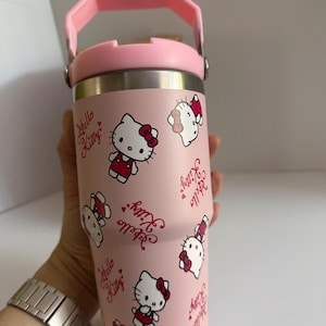 May include: A pink insulated tumbler featuring a repeating pattern of Hello Kitty characters and text. The tumbler has a pink handle and lid, and a silver-colored body. The design includes Hello Kitty in a red dress and bow.