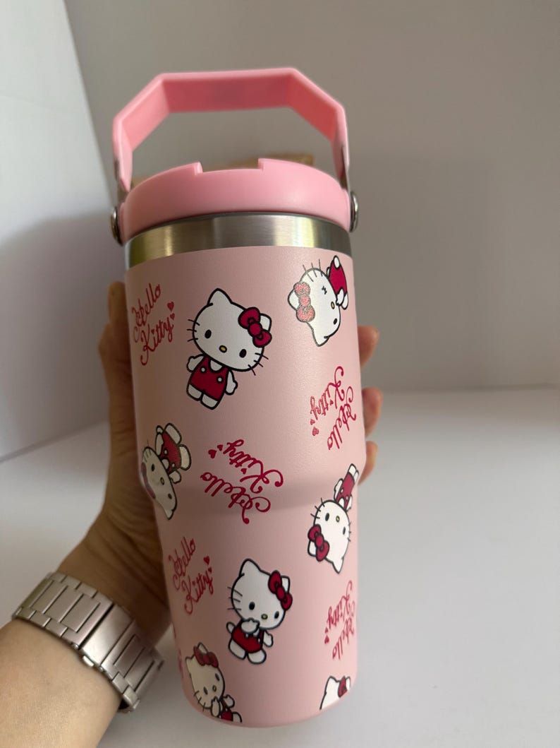 May include: A pink insulated tumbler featuring a repeating pattern of Hello Kitty characters and text. The tumbler has a pink handle and lid, and a silver-colored body. The design includes Hello Kitty in a red dress and bow.