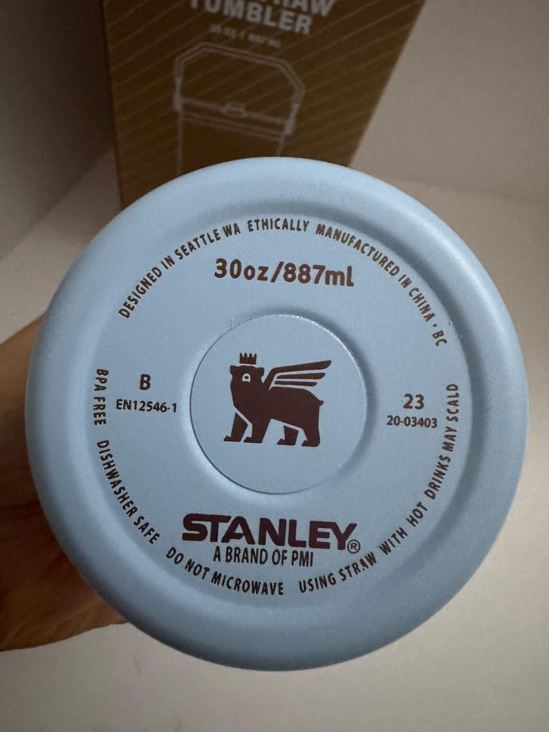 Puede incluir: Base de vaso Stanley azul claro con el texto "30oz/887ml". La base incluye el logotipo de Stanley, un oso con alas y corona, y el texto "BPA FREE DISHWASHER SAFE DO NOT MICROWAVE USING STRAW WITH HOT DRINKS MAY SCALD".