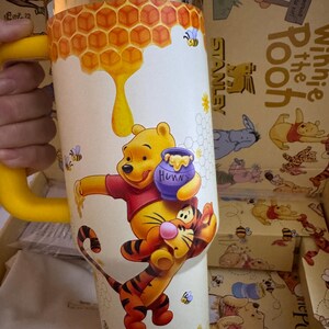 May include: A yellow and silver Stanley travel mug featuring Winnie the Pooh and Tigger. The mug has a yellow handle and a design of honeycombs, bees, and Winnie the Pooh holding a honey pot. The mug is in a box with Winnie the Pooh illustrations.