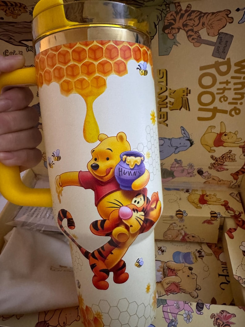 May include: A yellow and silver Stanley travel mug featuring Winnie the Pooh and Tigger. The mug has a yellow handle and a design of honeycombs, bees, and Winnie the Pooh holding a honey pot. The mug is in a box with Winnie the Pooh illustrations.