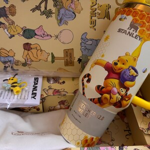 May include: A yellow and white Stanley Quencher 12.0 tumbler featuring Winnie the Pooh and Tigger. The tumbler has a yellow handle and a honeycomb design. A yellow bee-shaped accessory is also visible.