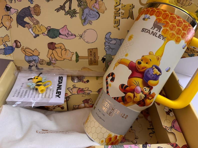 May include: A yellow and white Stanley Quencher 12.0 tumbler featuring Winnie the Pooh and Tigger. The tumbler has a yellow handle and a honeycomb design. A yellow bee-shaped accessory is also visible.