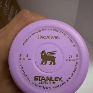 Puede incluir: Primer plano de una tapa de botella de agua Stanley de color lila claro. La tapa tiene el texto "30oz/887ml" y un logotipo de un oso con alas. Tambi&eacute;n se puede leer "Dishwasher Safe" y "Do Not Microwave".