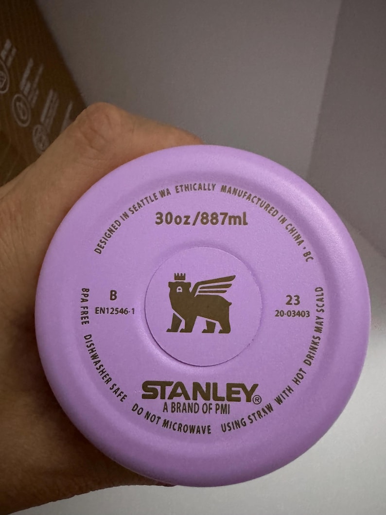 Puede incluir: Primer plano de una tapa de botella de agua Stanley de color lila claro. La tapa tiene el texto "30oz/887ml" y un logotipo de un oso con alas. Tambi&eacute;n se puede leer "Dishwasher Safe" y "Do Not Microwave".