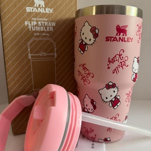 May include: A pink Stanley The Iceflow Flip Straw Tumbler, 20 oz, featuring Hello Kitty graphics. The tumbler has a pink lid and straw, and comes with its original box. The Stanley logo is visible on the tumbler and box.