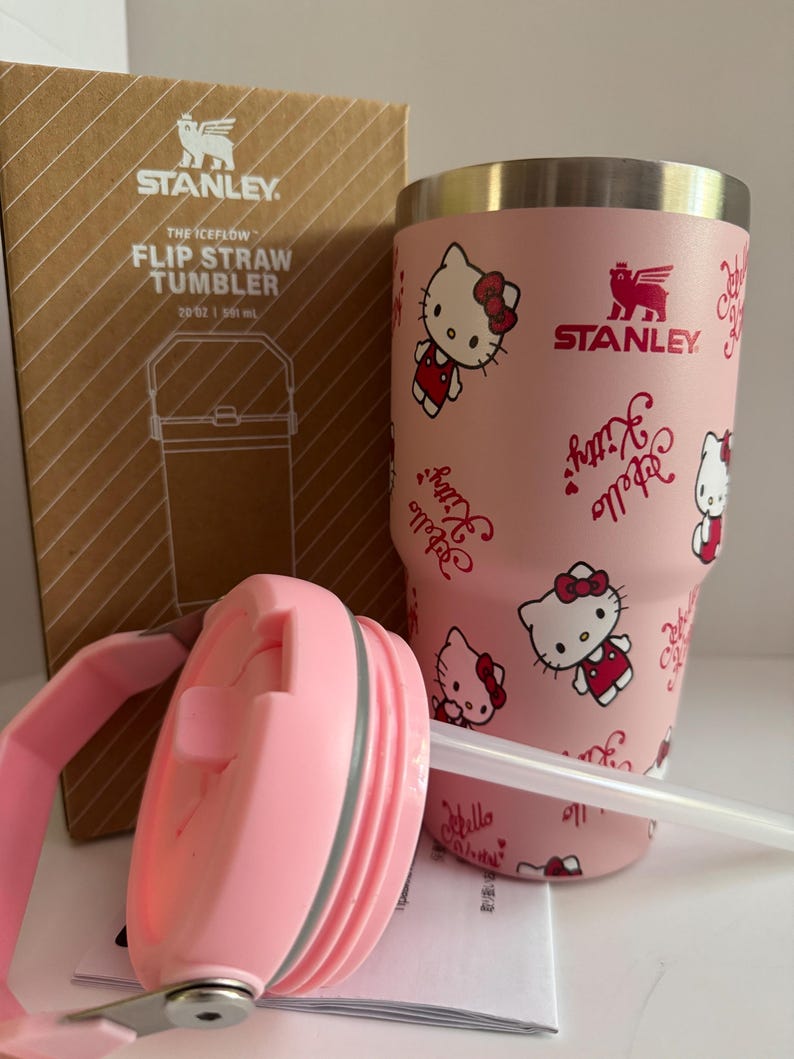 May include: A pink Stanley The Iceflow Flip Straw Tumbler, 20 oz, featuring Hello Kitty graphics. The tumbler has a pink lid and straw, and comes with its original box. The Stanley logo is visible on the tumbler and box.